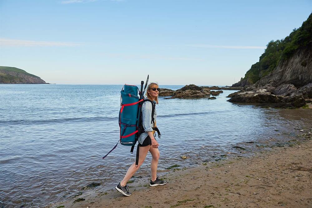 woman walking along a beach carrying a paddleboard bag