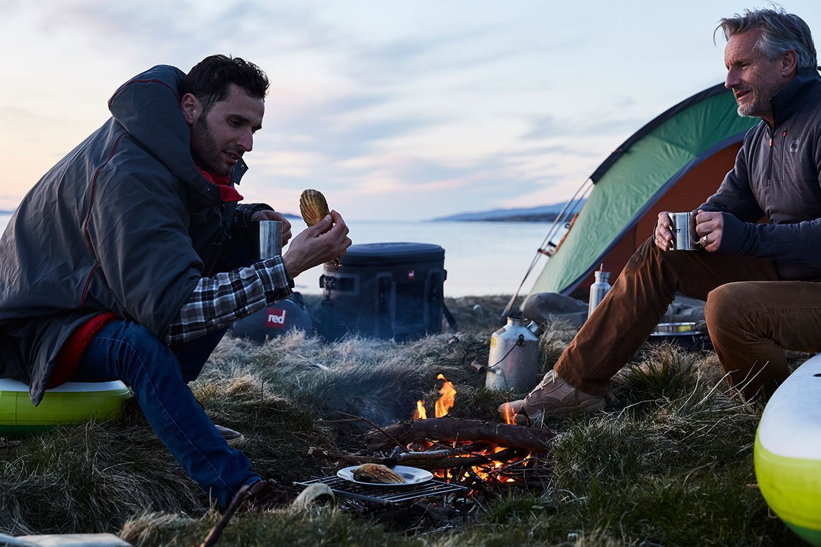 2 Men Enjoying Food By A Campfire On A Beach.